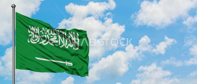 علم المملكة العربية السعودية يرفرف في مهب الريح أمام سماء زرقاء ملبدة بالغيوم البيضاء. مفهوم الدبلوماسية، العلاقات الدولية.