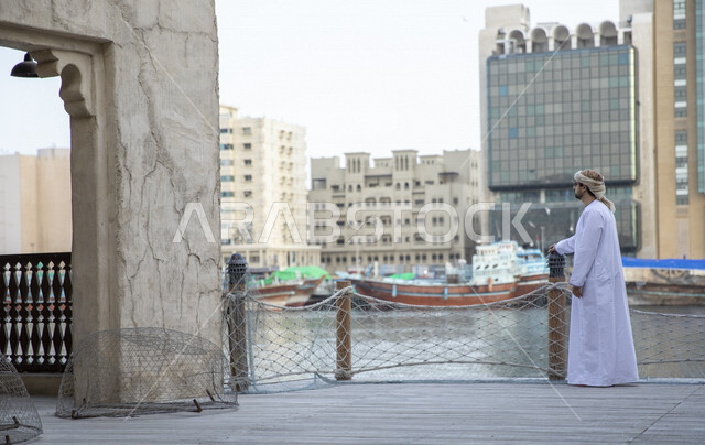 رجل عربي خليجي عماني يقف امام خور دبي في الإمارات العربية المتحدة، المدينة القديمة بدبي، القوارب الخشبية، الابراج السكنية، ميناء سفن، البيوت القديمة، معالم دبي المشهورة، صورة الجسم بالطول الكامل، السياحة في الإمارات