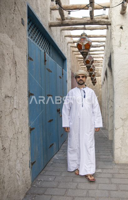 رجل عربي خليجي عماني يرتدي النظارات الشمسية ويتجول في شوارع حي السيف بدبي في الإمارات العربية المتحدة، احياء المدينة القديمة بدبي، التجول في احياء دبي، البيوت الشعبية التاريخية القديمة، معالم دبي المشهورة، صورة الجسم بالطول الكامل، السياحة في الامارات