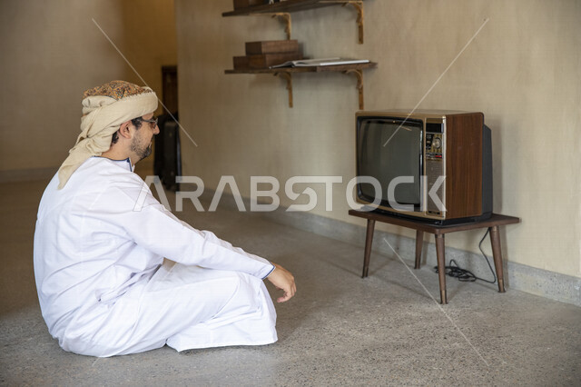 رجل عربي خليجي عماني يرتدي النظارات الشمسية ويجلس امام تلفزيون قديم داخل ارجاء البيت القديمة، تلفزيون اثري قديم، البيوت الشعبية القديمة في حي السيف بدبي، البيوت الاثرية، منازل تاريخية، السياحة في الإمارات