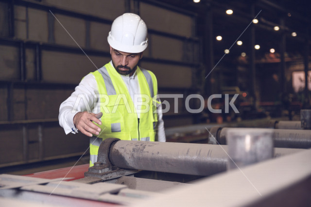 مهندس ومشرف عربي خليجي سعودي يرتدي خوذة و سترة الحماية الخاصة بالعمل في مصنع الحديد ، يتحقق من جودة الحديد المنتج ، مفهوم الهندسة و الصناعة