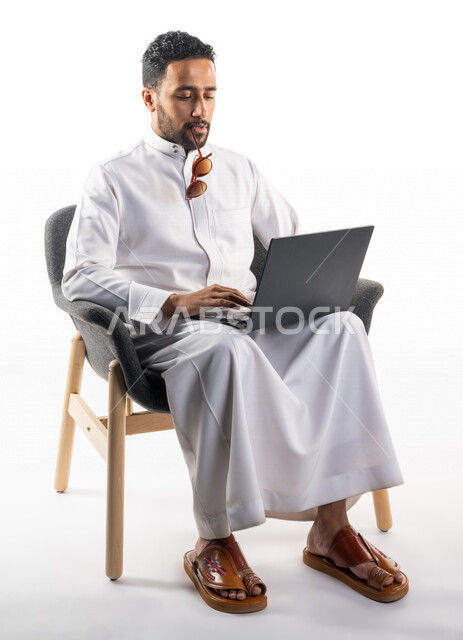 بورتريه لرجل عربي خليجي سعودي يجلس على كرسي يحمل بيده جهاز الحاسوب، اجراء مكالمات فيديو، تواصل اجتماعي، محادثات ودردشة، التواصل مع الأصدقاء، اجتماع عمل عن بعد، بث فيديو مباشر، خلفية بيضاء