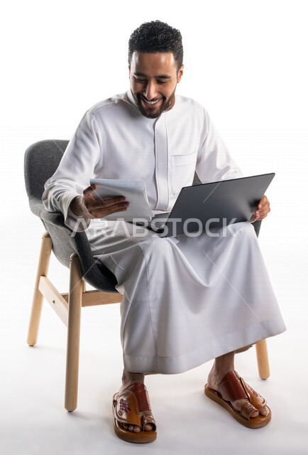 بورتريه لرجل عربي خليجي سعودي يجلس على كرسي يحمل بيده جهاز الحاسوب وأوراق عمل، إنجاز مهام العمل، تواصل اجتماعي، اجتماع عمل عن بعد، خلفية بيضاء