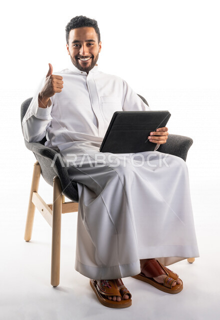 بورتريه لرجل عربي خليجي سعودي يجلس على كرسي يحمل بيده جهاز الحاسوب، اجراء مكالمات فيديو، تواصل اجتماعي، محادثات ودردشة، التواصل مع الأصدقاء، اجتماع عمل عن بعد، بث فيديو مباشر، خلفية بيضاء