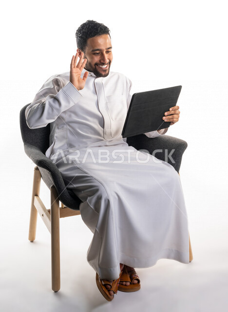 بورتريه لرجل عربي خليجي سعودي يجلس على كرسي يحمل بيده جهاز الحاسوب، اجراء مكالمات فيديو، تواصل اجتماعي، محادثات ودردشة، التواصل مع الأصدقاء، اجتماع عمل عن بعد، بث فيديو مباشر، خلفية بيضاء