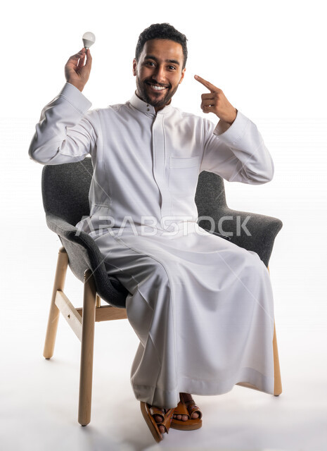 بورتريه لرجل سعودي خليجي يجلس على كرسي يحمل المصباح الضوئي بإيماءات وجه ويدين تدل على الذكاء ، أفكار إبداعية تطويرية ، مفهوم الفكرة مع الإبتكار والإلهام ، تحقيق الإنجازات والإبتكارات ، براءة الإختراع ، خلفية بيضاء ، المصباح الكهربائي الضوئي