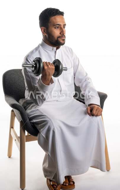 بورتريه لرجل عربي خليجي سعودي يجلس على كرسي يقوم بممارسة التمارين الرياضية بالدمبل ، نشاط بدني للمحافظة على الصحة و اللياقة الجسدية ، تقوية عضلات الجسم و بنائها ، تمارين رفع الأثقال (دمبلز)، رفع الأوزان الحديدية الثقيلة ، كمال الأجسام ، لياقة و رشاقة