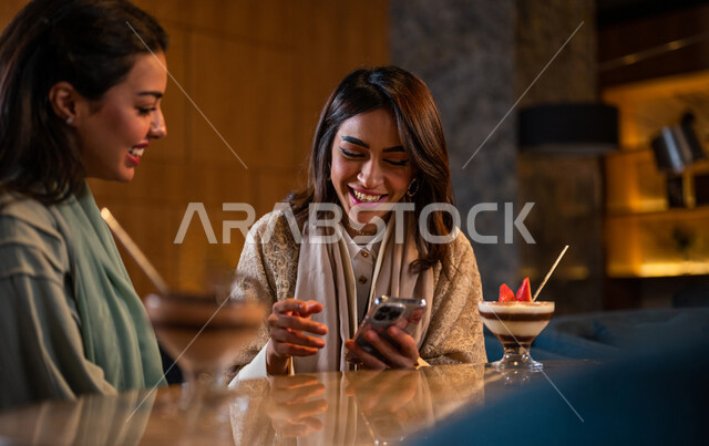 Saudi Arabian Gulf Arab female friends sitting in a café, using a mobile phone, online shopping, social relations between friends, using social media, spending fun times with friends, surfing the Internet, meeting and dating