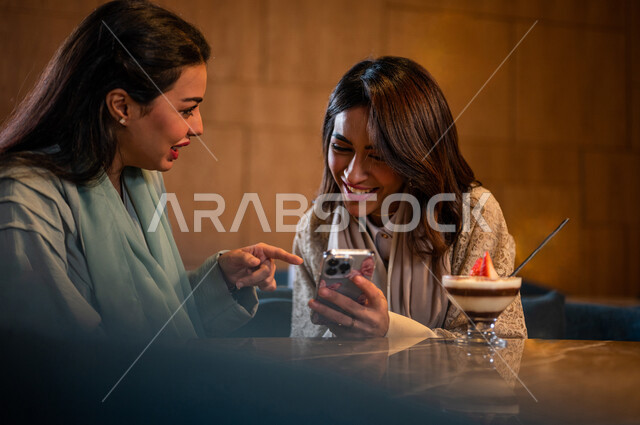 Saudi Arabian Gulf Arab female friends sitting in a café, using a mobile phone, online shopping, social relations between friends, using social media, spending fun times with friends, surfing the Internet, meeting and dating