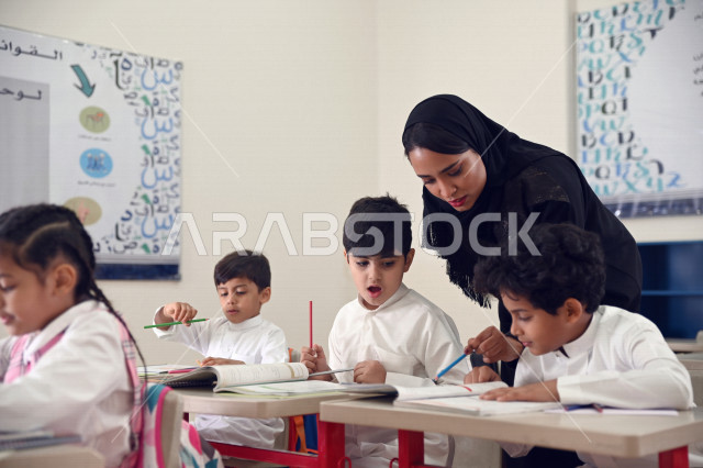 معلمة عربية سعودية خليجية تقوم بمراقبة الطلاب والطالبات ومساعدتهم بقراءة وحل المسائل , نشاطات مدرسية جماعية , أهداف التربية والتعليم بالمناهج المقررة للطلاب والطالبات في المملكة العربية السعودية .