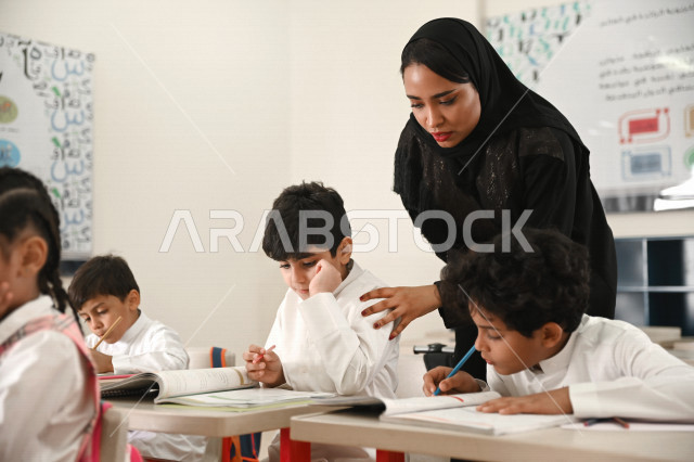 معلمة عربية سعودية خليجية تقوم بمراقبة الطلاب والطالبات ومساعدتهم بقراءة وحل المسائل , نشاطات مدرسية جماعية , أهداف التربية والتعليم بالمناهج المقررة للطلاب والطالبات في المملكة العربية السعودية .