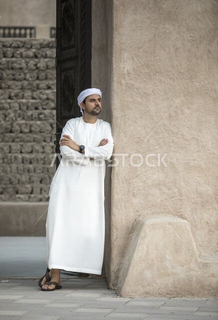 رجل عربي خليجي إماراتي، بالزي الإماراتي التقليدي، يقف بأيدي متقاطعة مع خلفية لأحياء دبي القديمة، البيوت الشعبية القديمة، معالم دبي التايخية الأثرية، الإمارات العربية المتحدة ، صورة بالطول الكامل، كامل الجسم