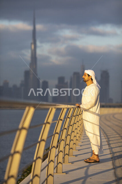 رجل عربي خليجي إماراتي، بالزي الإماراتي التقليدي، خلفية ضبابية لأبراج دبي، أبراج وناطحات السحاب على ساحل الخليج العربي، برج خلفية، معالم دبي السياحية المشهورة، الإمارات العربية المتحدة، ممشى كورنيش دبي، الواجهة البحرية في دبي، صورة بالطول الكامل