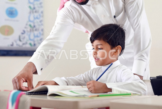 معلم عربي سعودي خليجي يقوم بمساعدة الطلاب لحل الأسئلة الموجودة في الكتب المدرسية , حل الواجبات المدرسية في الفصل , المقررات والمناهج التعليمية الحديثة والمطورة في المملكة العربية السعودية .