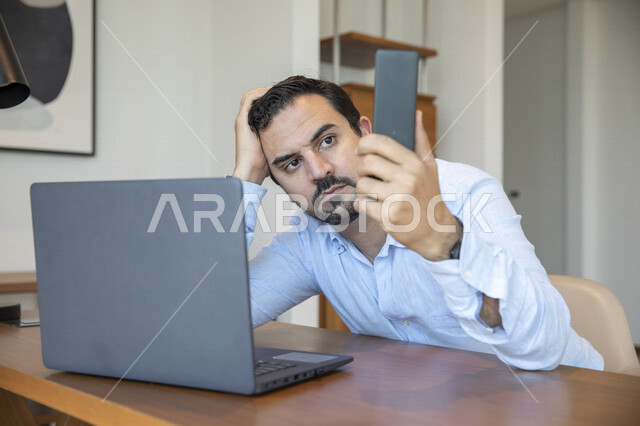 رجل عربي يحمل بيده لوح تقني ، إيماءات تدل على الإحباط ، إستخدام جهاز الحاسوب المحمول، العمل عن بعد ، تصفح الإنترنت ، الشعور بالضجر والملل