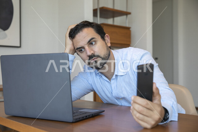 رجل عربي يحمل بيده لوح تقني ، إيماءات تدل على الإحباط ، إستخدام جهاز الحاسوب المحمول، العمل عن بعد ، تصفح الإنترنت ، الشعور بالضجر والملل