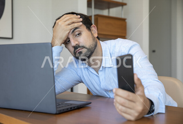 رجل عربي يحمل بيده لوح تقني ، إيماءات تدل على الإحباط ، إستخدام جهاز الحاسوب المحمول، العمل عن بعد ، تصفح الإنترنت ، الشعور بالضجر والملل