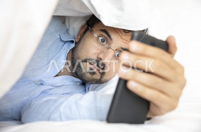 An Arab man lying on the bed, wearing medical glasses for low vision, using a mobile phone, gestures of astonishment and amazement, shock to see something, watching programs, surfing the Internet, using social media