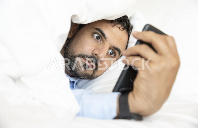 رجل عربي مستلقي على فراش النوم ، إستخدام الهاتف المحمول ،  إيماءات تدل الدهشة والذهول ، الصدمة لرؤية شيء ما ، مشاهدة البرامج ، تصفح الإنترنت ، إستخدام وسائل التواصل الإجتماعي