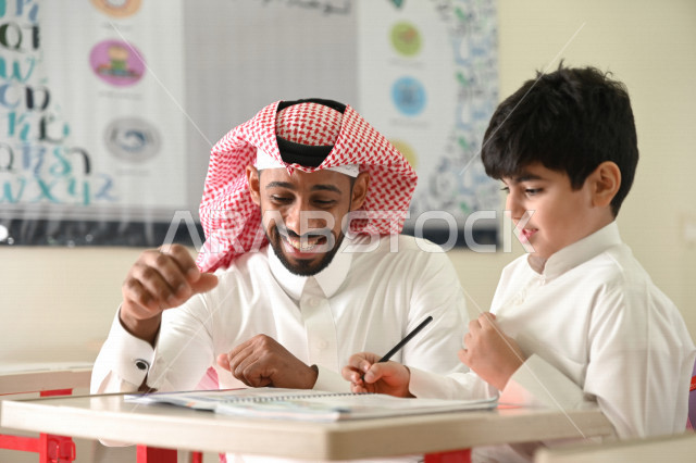 معلم عربي سعودي خليجي يقوم بمساعدة الطلاب لحل الأسئلة الموجودة في الكتب المدرسية , حل الواجبات المدرسية في الفصل , المقررات والمناهج التعليمية الحديثة والمطورة في المملكة العربية السعودية .