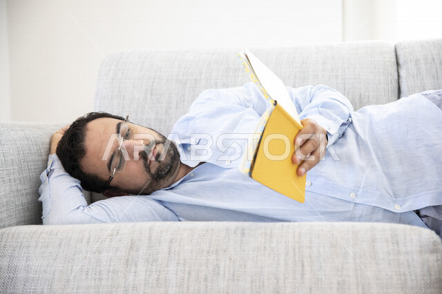 رجل عربي مستلقي على الكنبه ، يحمل بيده كتاب ، القراءة والتثقف ، قضاء أوقات الفراغ بالقراءة ، دفتر ملاحظات أصفر ، ضعف البصر ، قراءة الملاحظات ، الرؤية من خلال النظارة الطبية