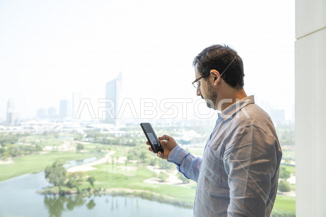 رجل عربي يرتدي النظارات الطبية ، يقف أمام النافذة ، يقوم بإستخدام الهاتف المحمول ، تصفح الإنترنت ، التواصل مع الأصدقاء ، العمل عن بعد ، جلسة عمل ، التسويق الإلكتروني