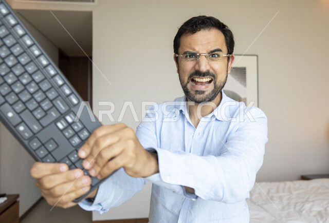 رجل عربي يرتدي النظارات الطبية لضعف النظر، يحمل بيده لوحة المفاتيح بإيماءات تدل على الغصب والتعصب ، مفهوم التوتر و الضغط العصبي والنفسي ، الشعور بالضجر  و الإنزعاج