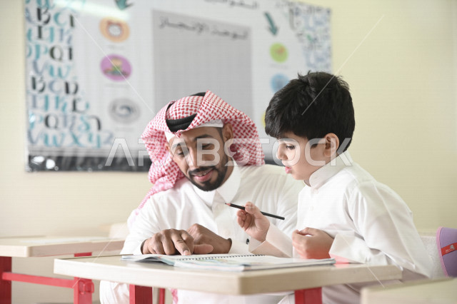 معلم عربي سعودي خليجي يقوم بمساعدة الطلاب لحل الأسئلة الموجودة في الكتب المدرسية , حل الواجبات المدرسية في الفصل , المقررات والمناهج التعليمية الحديثة والمطورة في المملكة العربية السعودية .