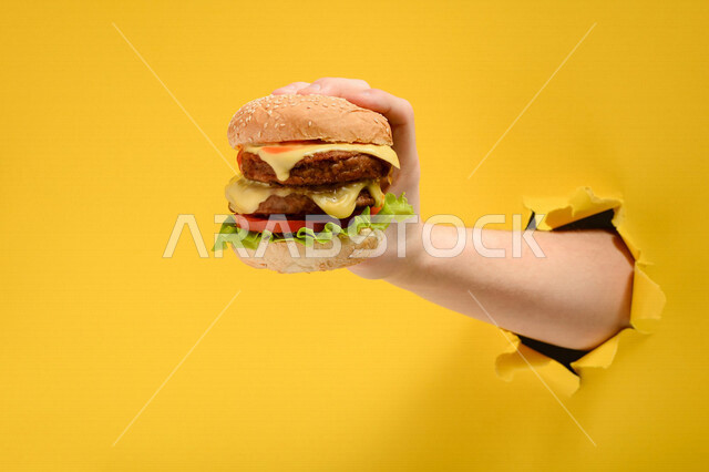 يد تمسك ساندوتش البرجر اللذيذ من خلال ثقب في جدار ورقي اصفر، اعلانات وعروض، الوجبات السريعة، وصفات طعام لذيذة، وجبة الهمبرجر اللذيذة، مطاعم ومأكولات، مطاعم عربية، خلفية صفراء