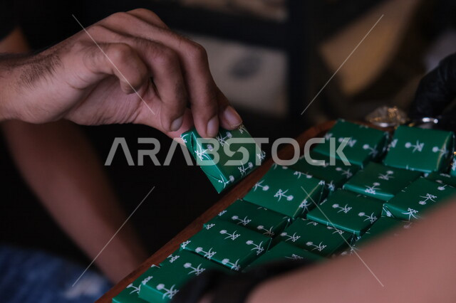 صورة مقربة ليد رجل عربي خليجي سعودي يمسك حبة الشوكلاتة، قطع الشوكلاتة، شوكلاتة اليوم الوطني السعودي، الاحتفال بيوم الاستقلال السعودي، اليوم الوطني السعودي