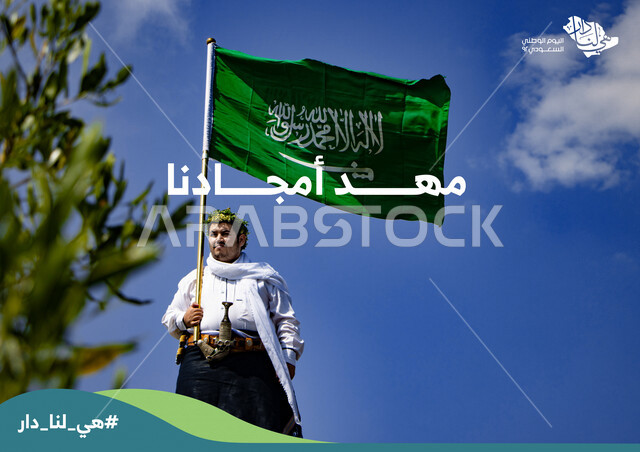 شاب عربي خليجي سعودي ، يحمل بيده علم السعودية من فوق جبال الحشر في محافظة الداير في منطقة جازان بالمملكة العربية السعودية في وضع النهار ، هوية اليوم الوطني السعودي 92 ، العيد الوطني للمملكة، هي لنا دار، شعار مهد أمجادنا