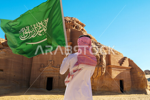 زاوية من الخلف لرجل عربي خليجي سعودي يحمل علم السعودية، الجمال التاريخي لمدائن صالح في محافظة العلا بالمملكة العربية السعودية، معالم تاريخية وأثرية قديمة في بلدة العلا القديمة، التكوينات الصخرية، هوية اليوم الوطني السعودي 92 ، رؤية السعودية 2030