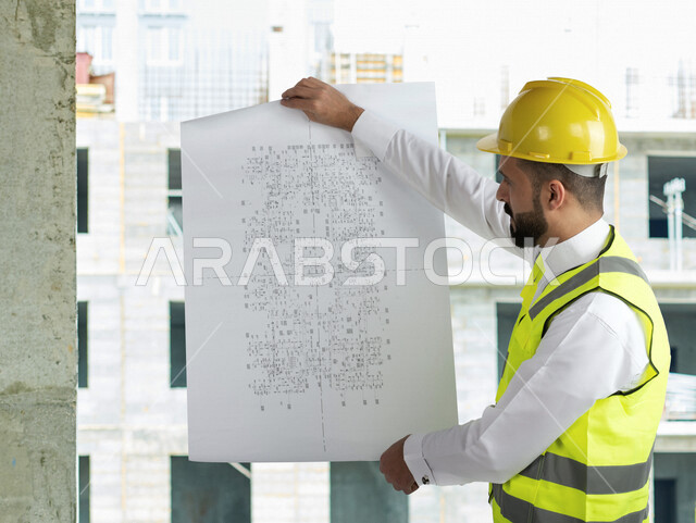 A Saudi Arabian Gulf architect wearing a work hat and protective suit, holding in his hand the construction plan, project follow-up and work audit, supervision and control of engineering projects, architecture, construction projects background.