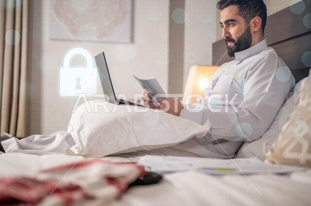  رجل عربي خليجي سعودي يستخدم الحاسوب المحمول في غرفة الفندق ، أيقونة قفل الأمان مضيئة بتقنية الهولوجرام ثلاثية الأبعاد ، الحماية و الأمان ، الحفاظ على البيانات الشخصية للمستخدمين الدخول الامن، مفهوم أمن تكنولوجيا المعلومات