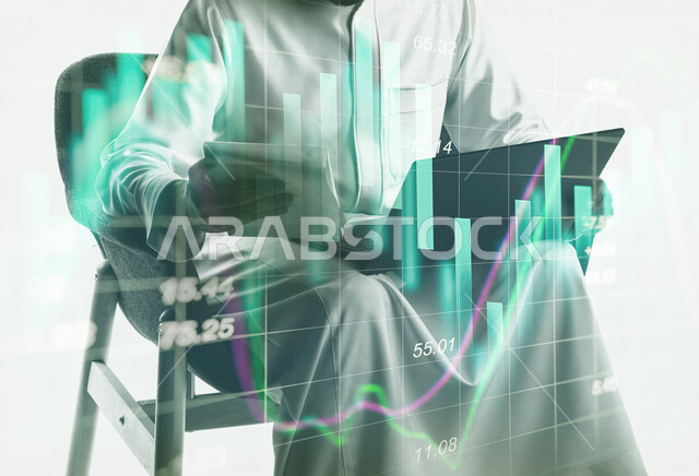 رجل أعمال عربي سعودي خليجي يجلس على كرسي ويقوم بالعمل على اللابتوب المحمول ،إستخدام أجهزة التقنية الحديثة لإنجاز المهام , تطوير وانشاء المشاريع الجديدة , رسومات البورصة بتقنية الهولوجرام . 