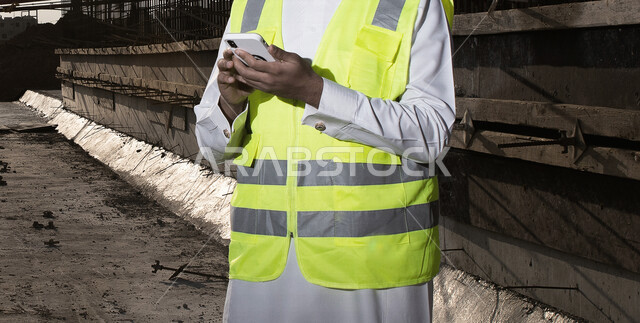 مهندس معماري عربي خليجي سعودي يرتدي سترة الوقايه الخاصه بالعمل، يستخدم الهاتف المحمول, متابعة المشروع وتدقيق العمل, ادارة اعمال البناء ، اشراف ومتابعة، الرقابه على المشاريع, هندسه معماريه