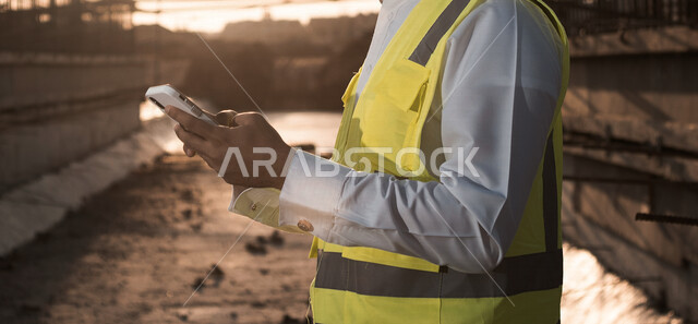 مهندس معماري عربي خليجي سعودي يرتدي سترة الوقايه الخاصه بالعمل، يستخدم الهاتف المحمول, متابعة المشروع وتدقيق العمل, ادارة اعمال البناء ، اشراف ومتابعة، الرقابه على المشاريع, هندسه معماريه