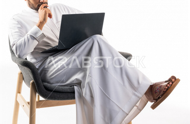 بورتريه لرجل عربي خليجي سعودي يجلس على كرسي يحمل بيده جهاز الحاسوب، اجراء مكالمات فيديو، تواصل اجتماعي، محادثات ودردشة، التواصل مع الأصدقاء، اجتماع عمل عن بعد، بث فيديو مباشر، خلفية بيضاء