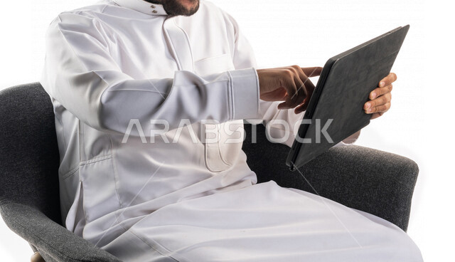 Portrait of a Saudi Arabian Gulf man sitting on a chair holding a technical device in his hand, making video calls, social communication, conversations and chatting, communicating with friends, remote work meeting, live video broadcasting, white backgroun