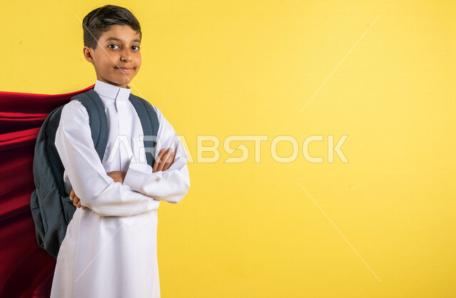 بورتريه لطالب عربي خليجي سعودي يرتدي الزي المدرسي، حقيبة الظهر المدرسية، أيدي متقاطعة، الإستعداد للعام الدراسي الجديد، مرحلة دراسية، التعليم في السعودية، الوقوف بثقة، ايماءات وجه ويدين تدل على الثقة، العودة إلى المدارس، خلفية صفراء