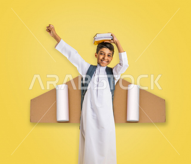 طالب عربي خليجي سعودي يرتدي الزي المدرسي ويضع الكتب المدرسية فوق رأسه، حقيبة الظهر المدرسية، إيماءات وجه ويدين تدل على السعادة، الإستعداد للعام الدراسي الجديد، التعليم في السعودية، العودة إلى المدارس، خلفية صفراء