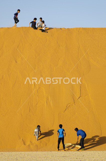 A group of children having fun on the sandy mountains in the desert in the Kingdom of Saudi Arabia, sand dunes, golden sand, playing and having fun, sand boarding, recreational activities in the desert, desert sand