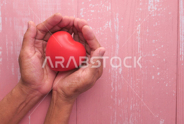 صورة مقربة ليد رجل يحمل مجسم قلب احمر صغير، مجسم على شكل رأس قلب، خلفية ملونة