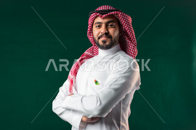 Karuma, Saudi Arabian Gulf Arab man looking at camera with crossed hands, wearing traditional Saudi Arabian Gulf dress, 2022 National Day logo on his chest, Saudi National Day celebration, Saudi National Day 92 ID, green background, Saudi founding day