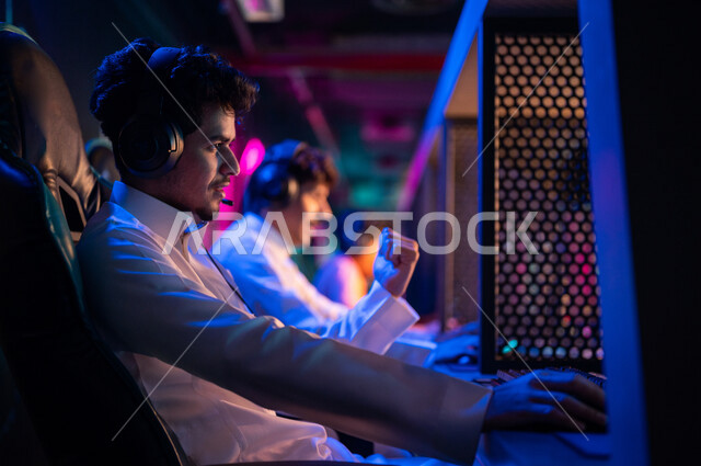 Young Arab Gulf Saudi teens in an electronic game store, wearing a headset and playing electronic video games, gestures indicating integration and interaction, recreational activities, computer entertainment, spending leisure time playing video games with