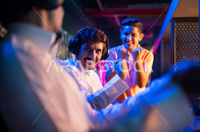 Young Arab Gulf Saudi teens in an electronic game store, wearing a headset and playing electronic video games, gestures indicating integration and interaction, recreational activities, computer entertainment, spending leisure time playing video games with