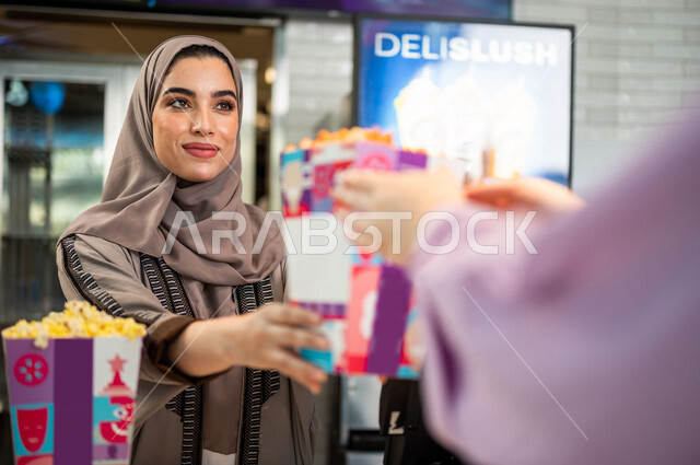  تصوير مقرب لإمرأه عربيه سعوديه خليجيه تبيع المأكولات والمشروبات في ردهة الإستقبال داخل السينما، تسليم الفشار للزبائن, الإستعداد للدخول الى صالة العرض، مشاهدة الأفلام والعروض المسرحية، سينما ومسرح، أنشطه ترفيهيه