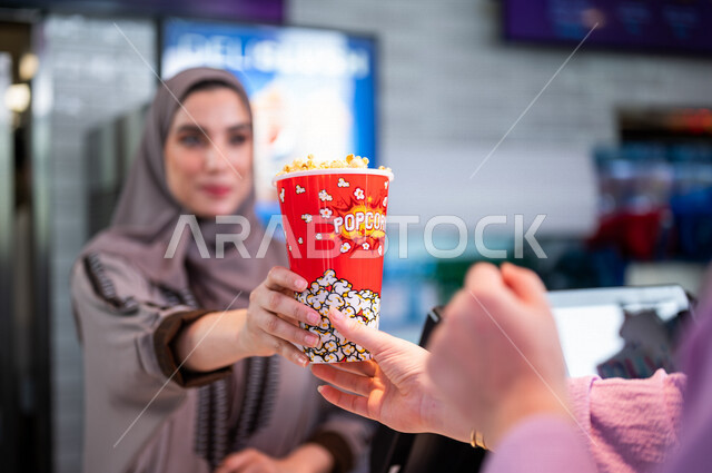  تصوير مقرب لإمرأه عربيه سعوديه خليجيه تبيع المأكولات والمشروبات في ردهة الإستقبال داخل السينما، تسليم الفشار للزبائن, الإستعداد للدخول الى صالة العرض، مشاهدة الأفلام والعروض المسرحية، سينما ومسرح، أنشطه ترفيهيه