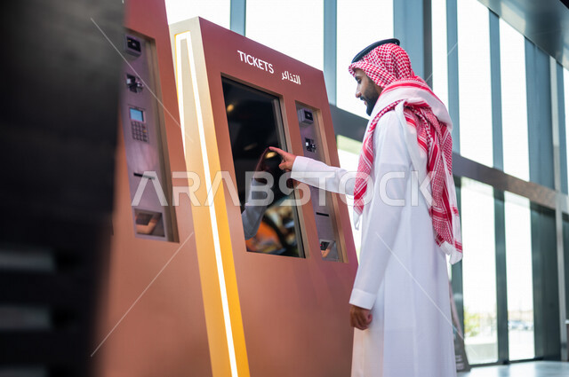 Photography from the back of a Saudi Arabian Gulf man wearing traditional Saudi dress booking a ticket to watch a movie in the cinema, watching movies and theatrical performances in the cinema, cinema and theater, entertainment activities