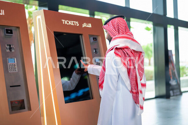  تصوير من الخلف لرجل عربي سعودي خليجي يرتدي الزي السعودي التقليدي يقوم بحجز تذكرة لمشاهدة فلم في صالة السينما، مشاهدة الأفلام والعروض المسرحية في السينما، سينما ومسرح، أنشطه ترفيهيه